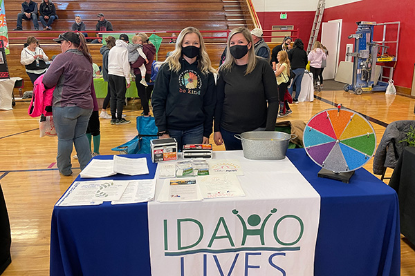Two women wearing masks stand behind an Idaho Lives information table. Click to enlarge.