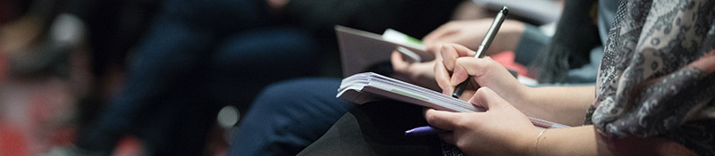Close up of a row of people sitting in a presentation, taking notes on notepads in their laps.