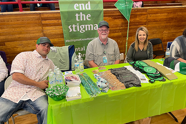 Two smiling men and a woman sitting behind an End the Stigma information table. Click to enlarge.